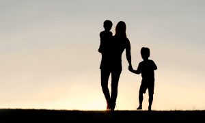 Silhouette of a young mother walking and lovingly holding hands with her happy little child, while holding his baby brother, outside in front of a sunset in the sky.